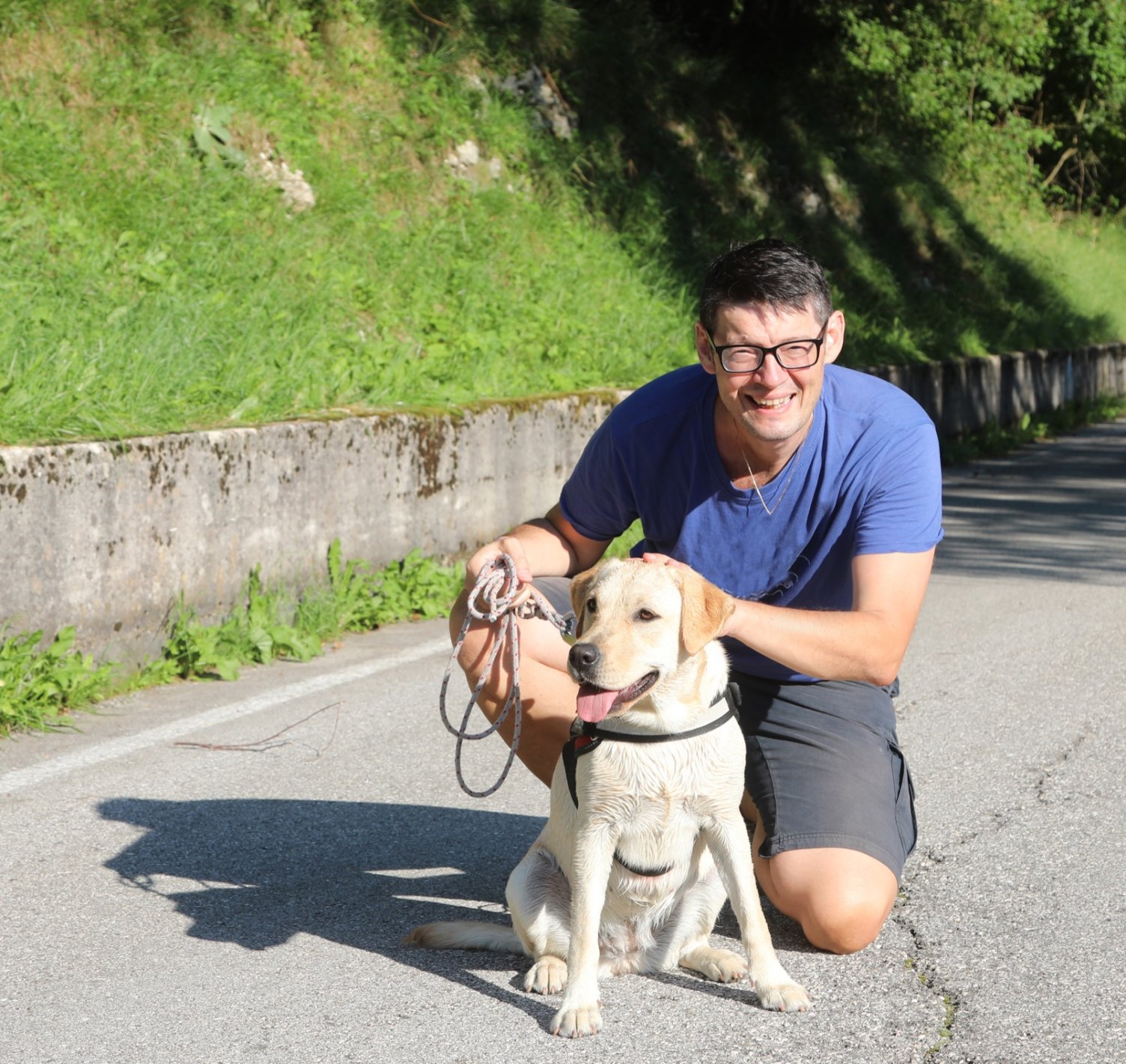 A man kneeling on a path beside his guide dog, both looking at the camera