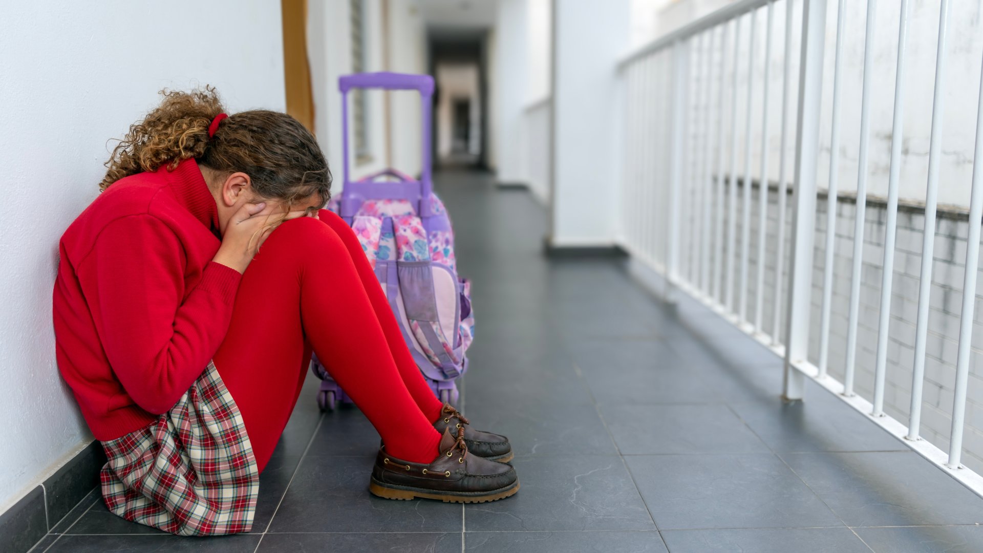 A child sitting on the floor of a school corridor with their head resting on their knees, a school bag beside them